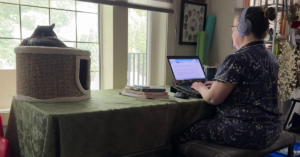 Woman sitting, working on laptop with cat sleeping next to her.